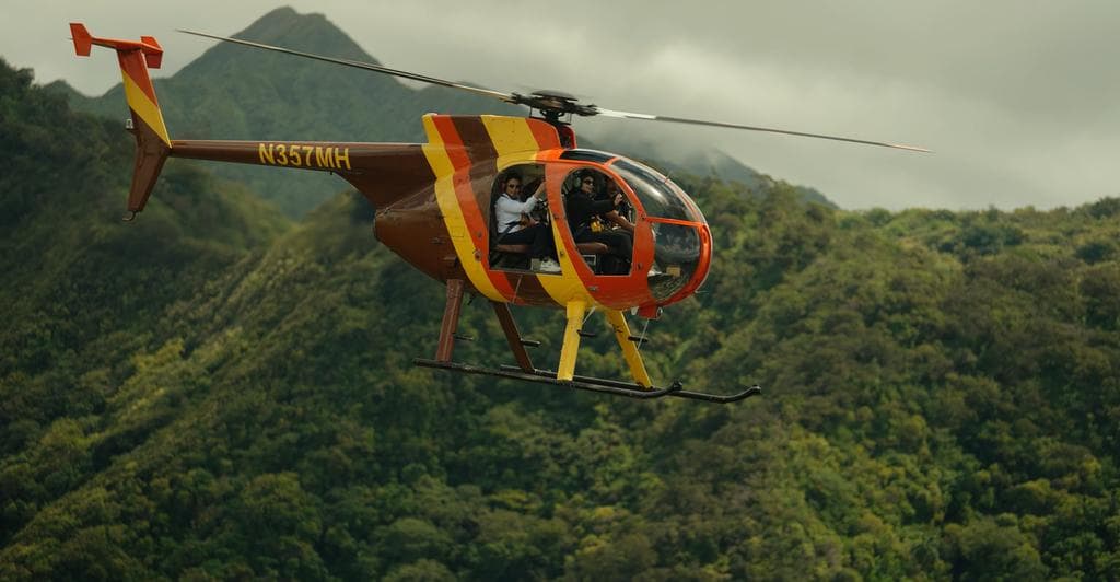 Hughes 500D helicopter in doors-off flight with green Oahu mountains behind