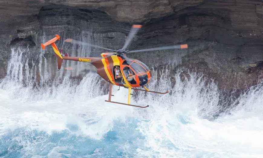 Doors-off helicopter hovering near an Oahu sea cliff with waves crashing below